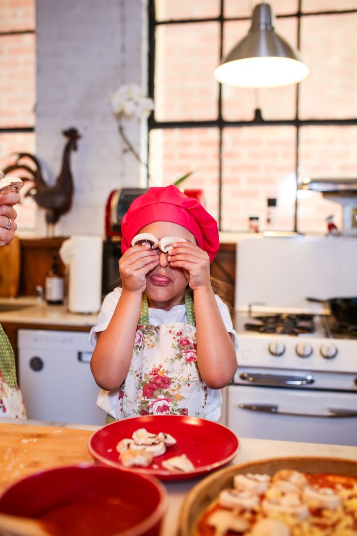 services-01 Young girl wearing a red chef hat and apron, playfully holding mushrooms in a cozy kitchen.